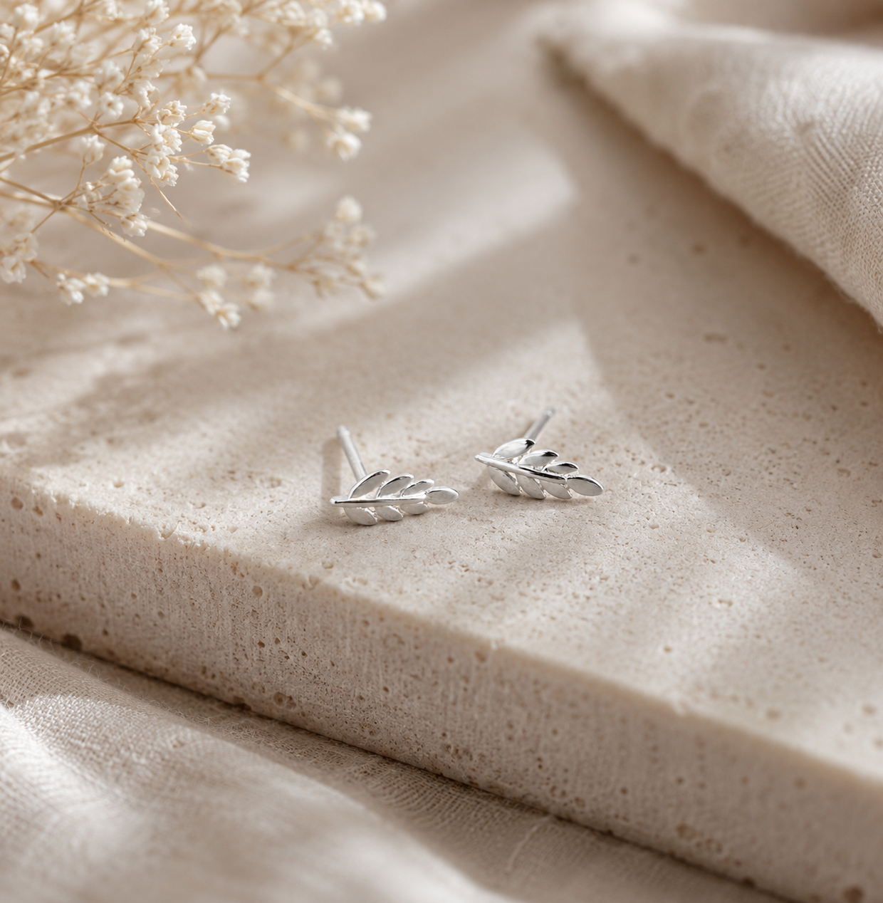 A pair of silver leaf-shaped stud earrings resting on a textured beige stone slab with soft neutral fabric and dried flowers in the background.