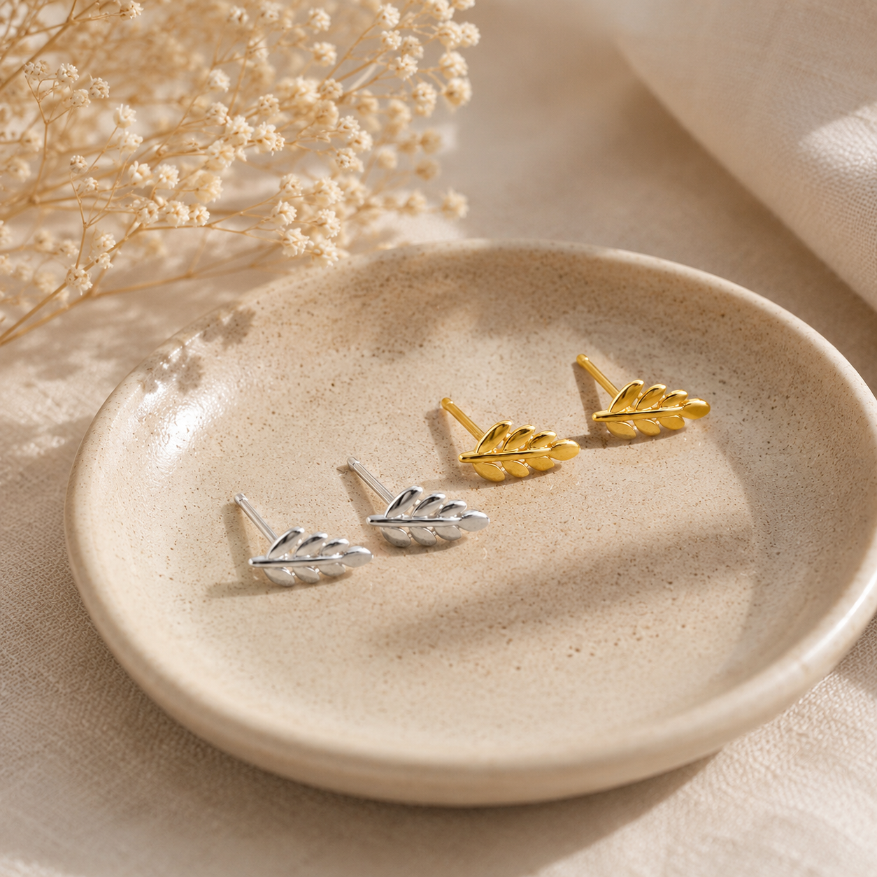 Three small leaf-shaped stud earrings, two silver-toned and one gold-toned, arranged on a round beige ceramic dish with dried flowers in the background.