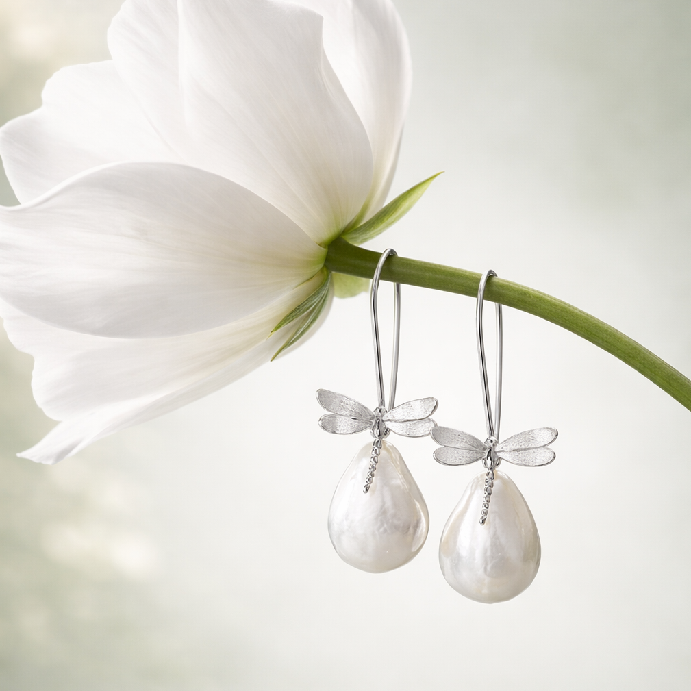 A pair of silver dragonfly drop earrings with teardrop-shaped white freshwater pearls, hanging from a green flower stem against a soft, out-of-focus white background.