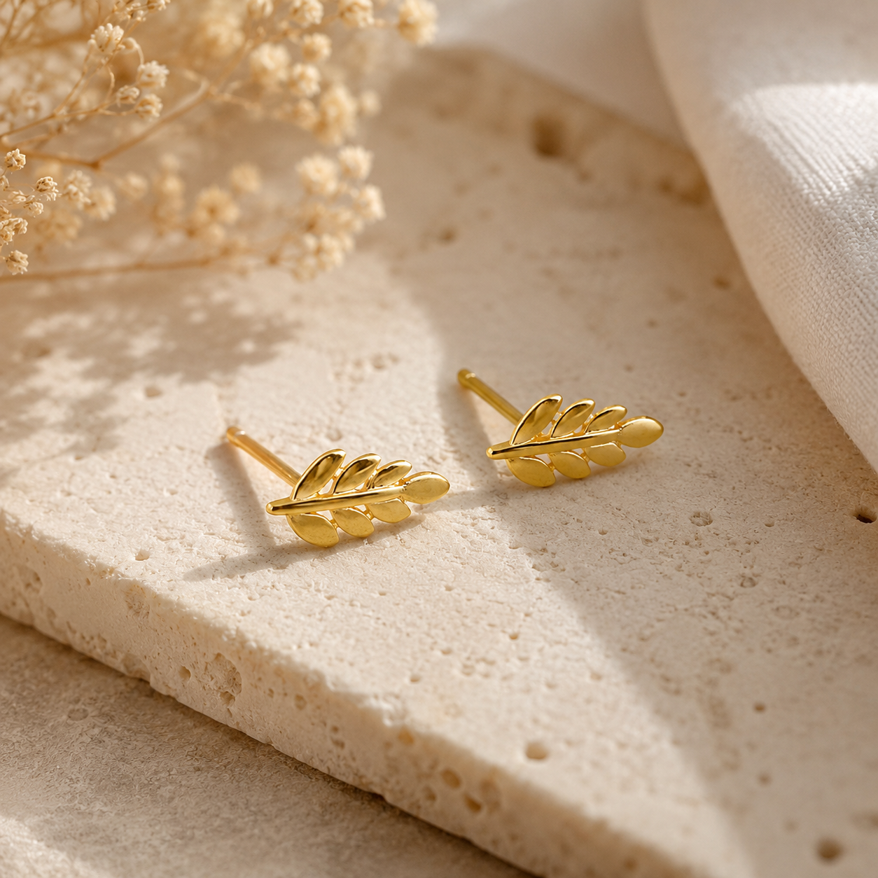 A pair of gold leaf-shaped stud earrings resting on a textured beige stone slab with dried flowers and soft white fabric in the background.