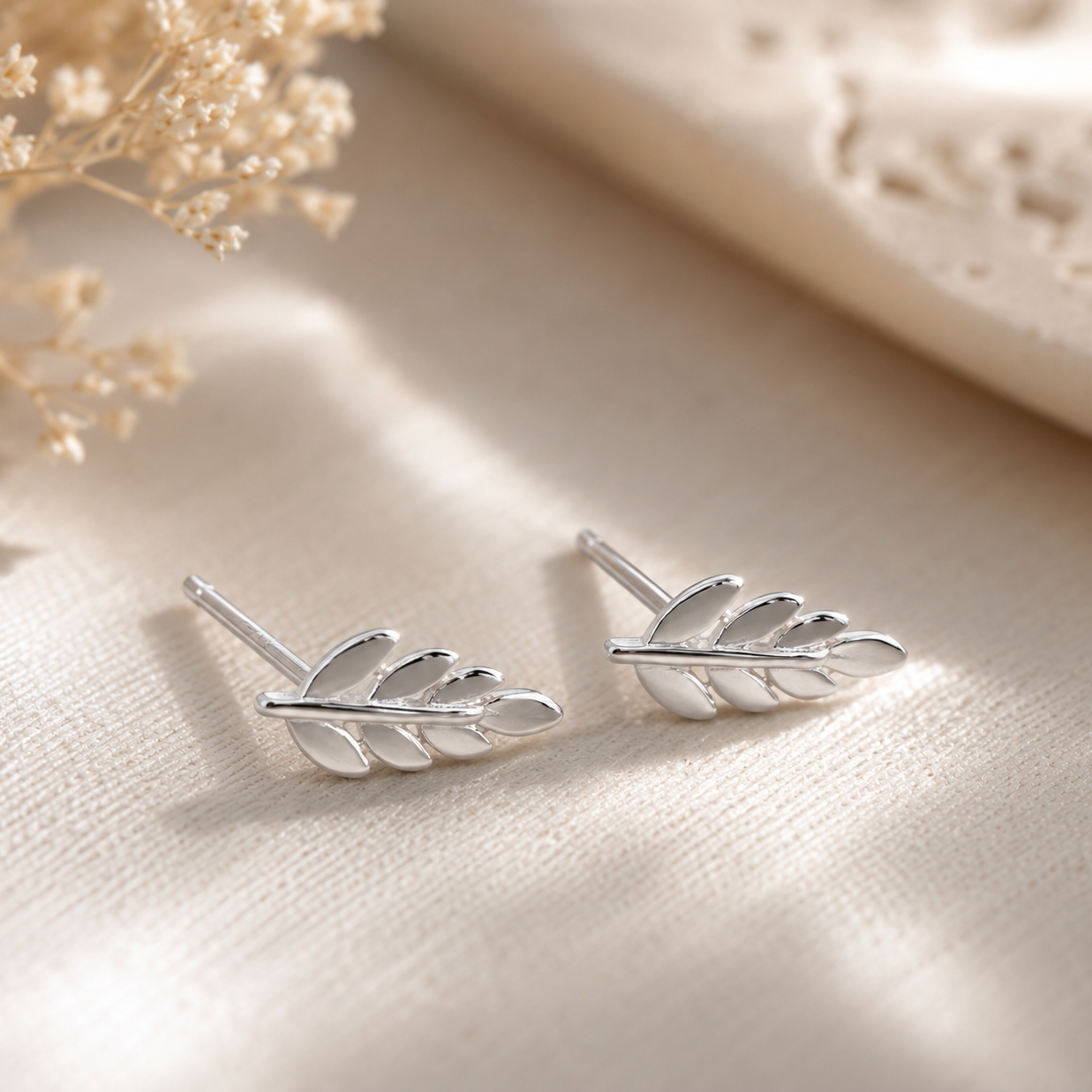 A pair of silver leaf-shaped stud earrings resting on a textured beige fabric surface with soft, warm light and dried flowers in the background.