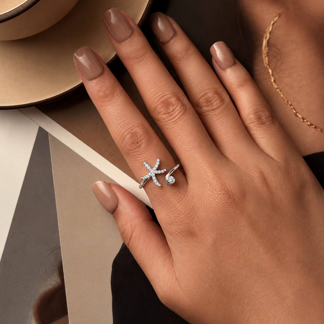 A close-up lifestyle shot of a model's hand wearing a silver-toned ring with a starburst and teardrop design. The hand has a neutral nude manicure, and the background features a beige hat, a gold chain necklace, and neutral geometric props.